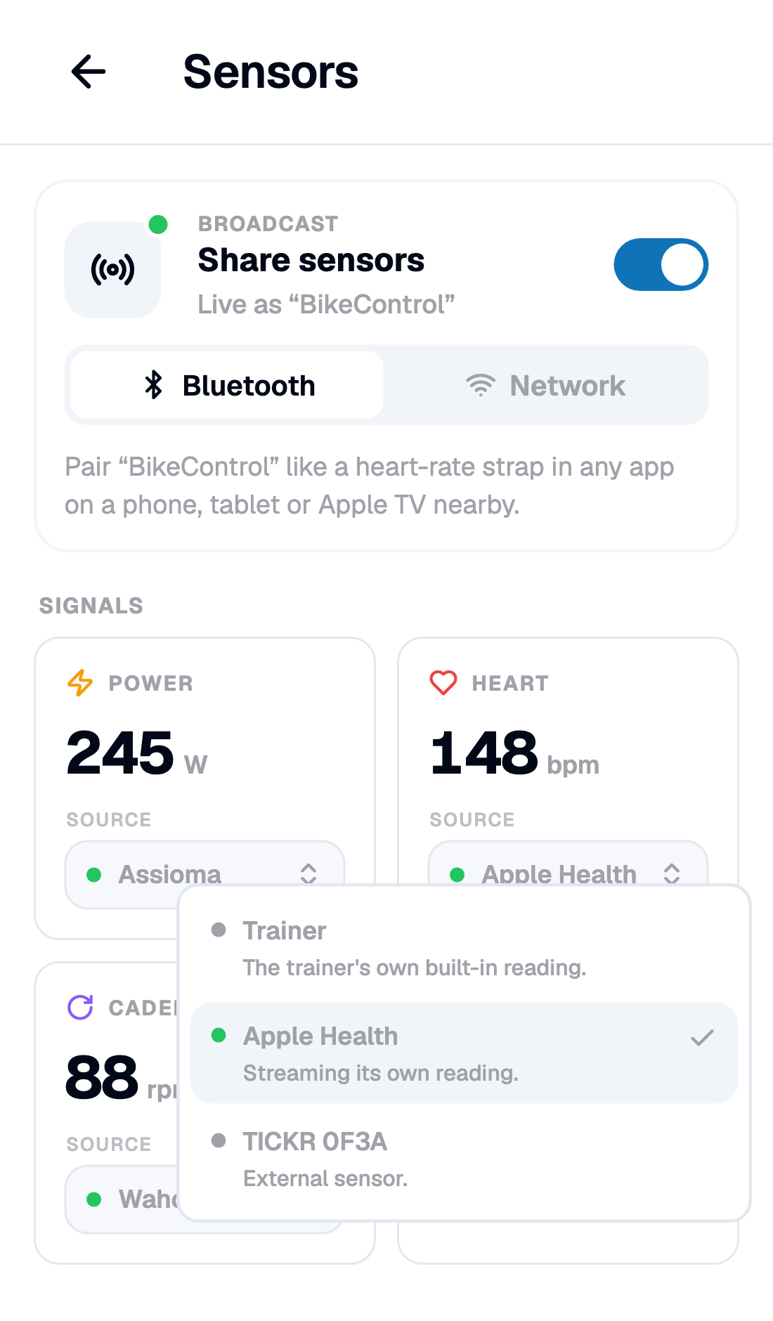 Apple Health sits in the list next to any strap. On iOS 26 it streams; below that, Health only shares what another workout is already recording The Sensors page with the Heart tile's Source list open over it: Trainer with "The trainer's own built-in reading", Apple Health ticked with "Streaming its own reading", and a TICKR strap listed as an external sensor; the tile reads 148 bpm